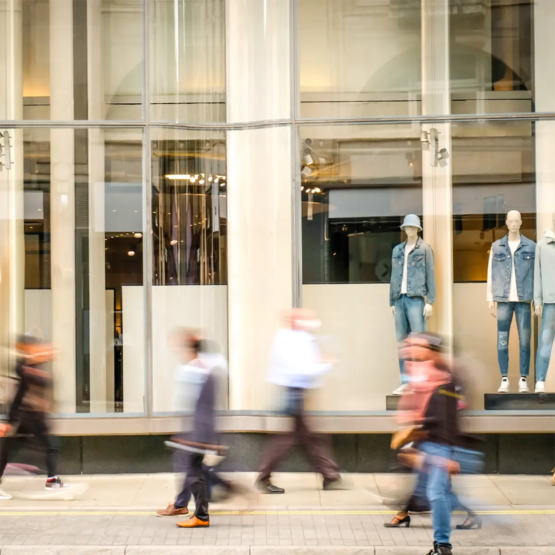 Blurry pedestrians walk past a shop window displaying mannequins dressed in denim jackets and jeans.