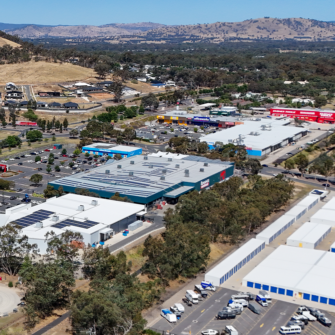 An aerial shot captures a sprawling commercial area with multiple large buildings, including a Bunnings Warehouse, all nestled amongst trees and hills.