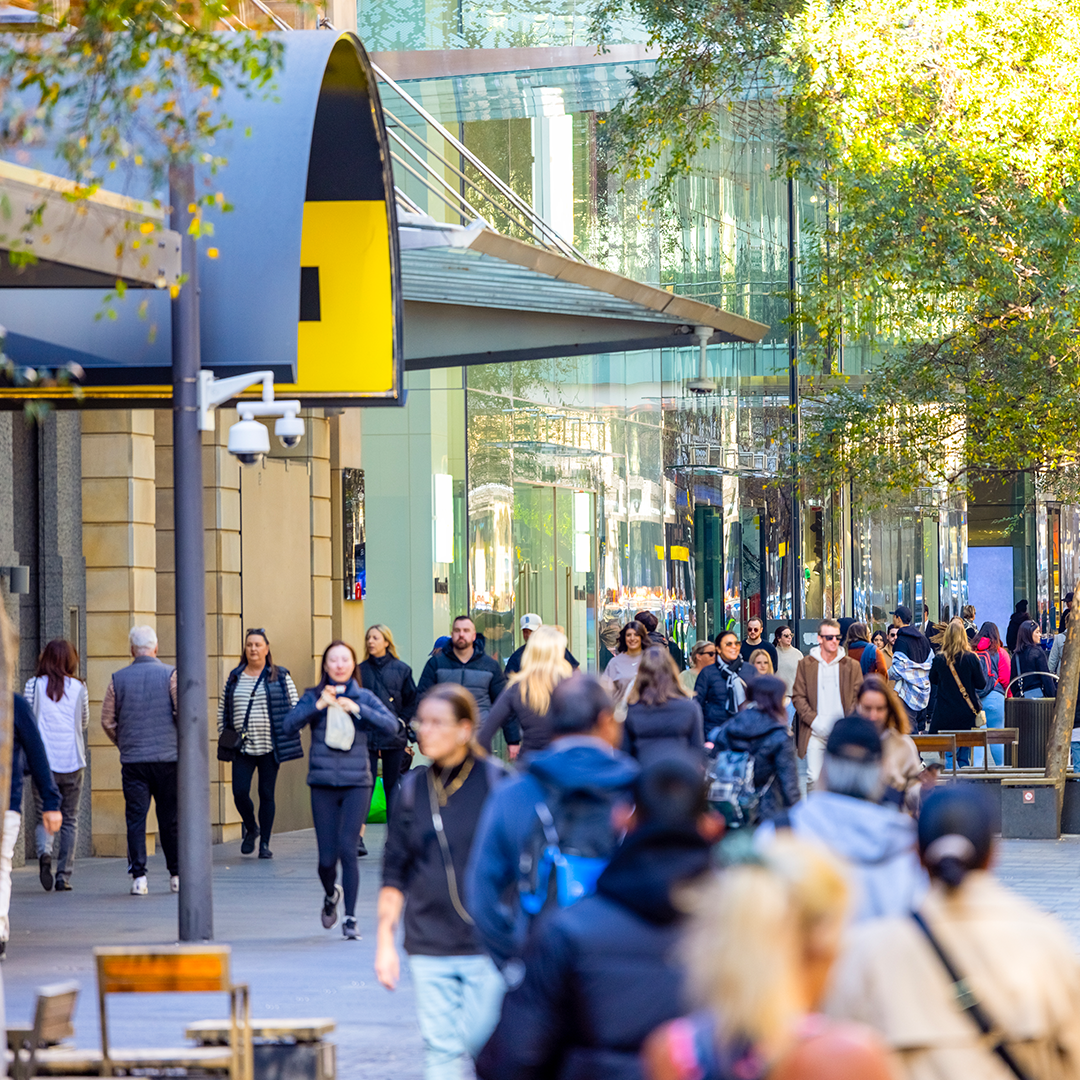 A bustling urban street scene shows numerous people walking along a footpath lined with modern glass-fronted buildings and trees, under a bright sky.