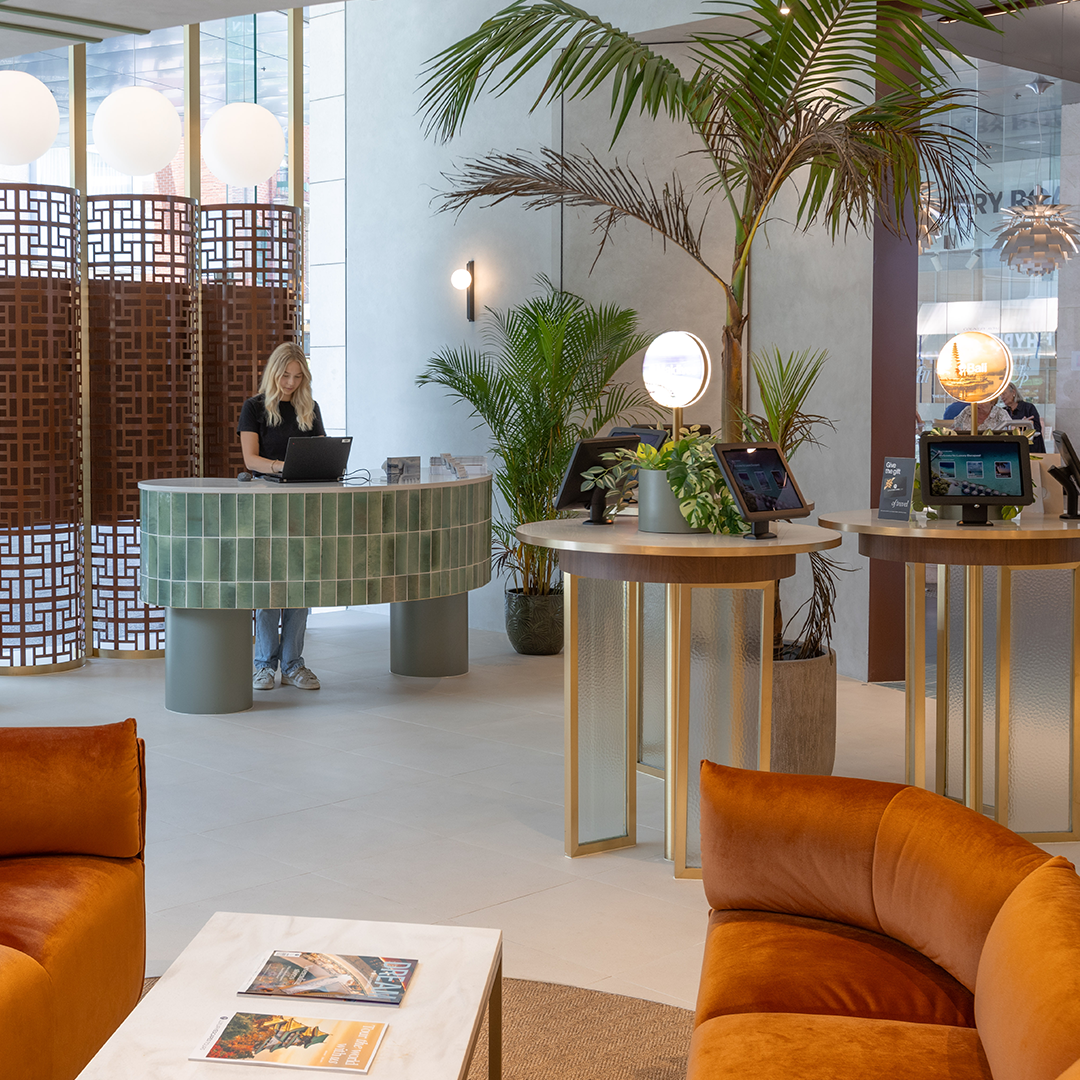 A modern, bright hotel lobby features a woman at a tiled reception desk, surrounded by potted plants, two round service tables with screens, and orange velvet armchairs in the foreground.