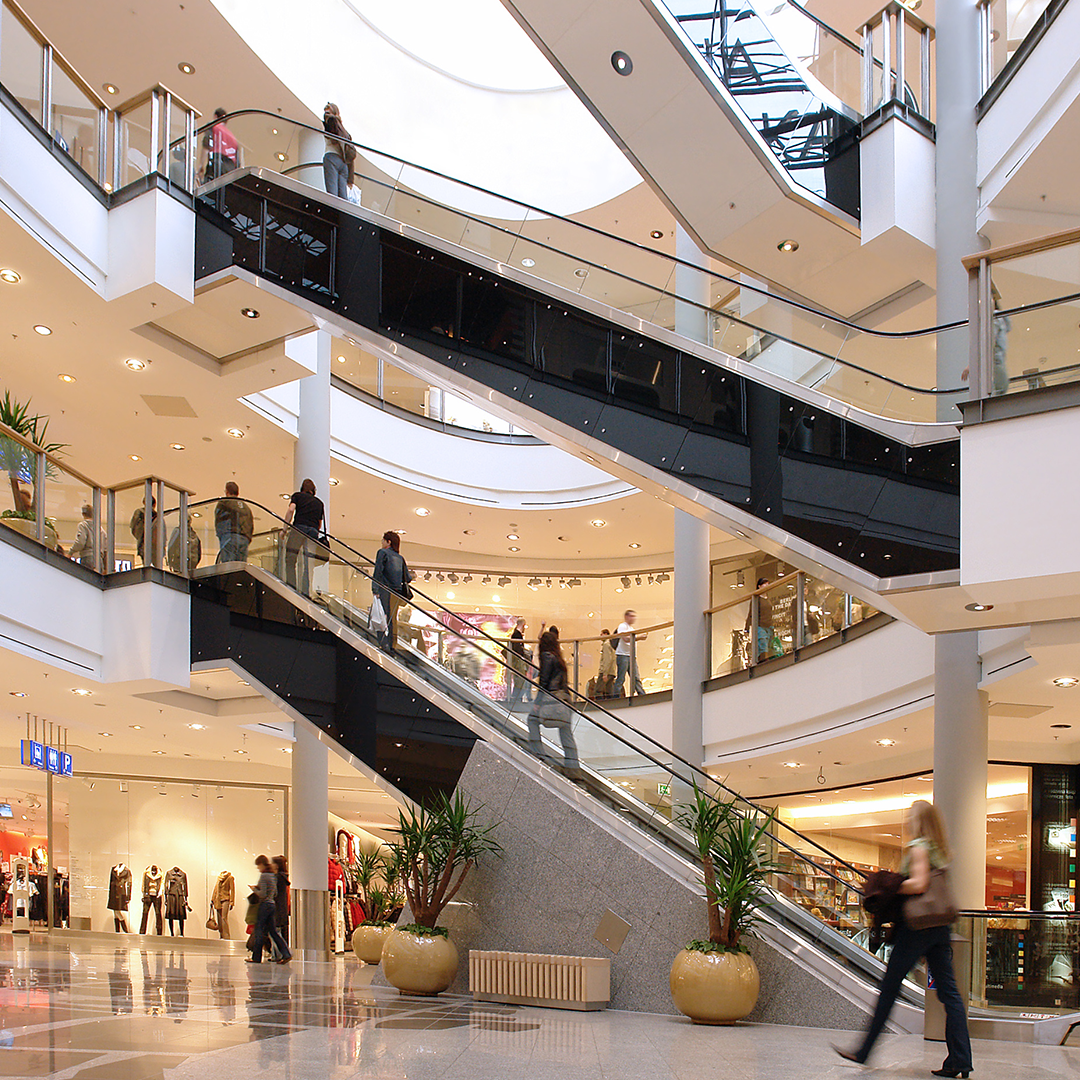 Shoppers move through a modern, multi-level shopping centre with escalators connecting the floors.