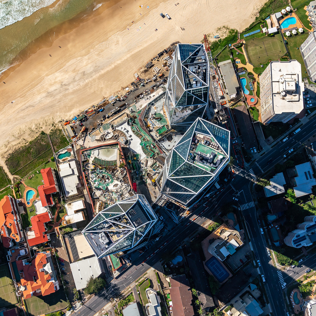 Aerial view of a coastal construction site featuring two distinctive, angular glass buildings under construction next to a beach.