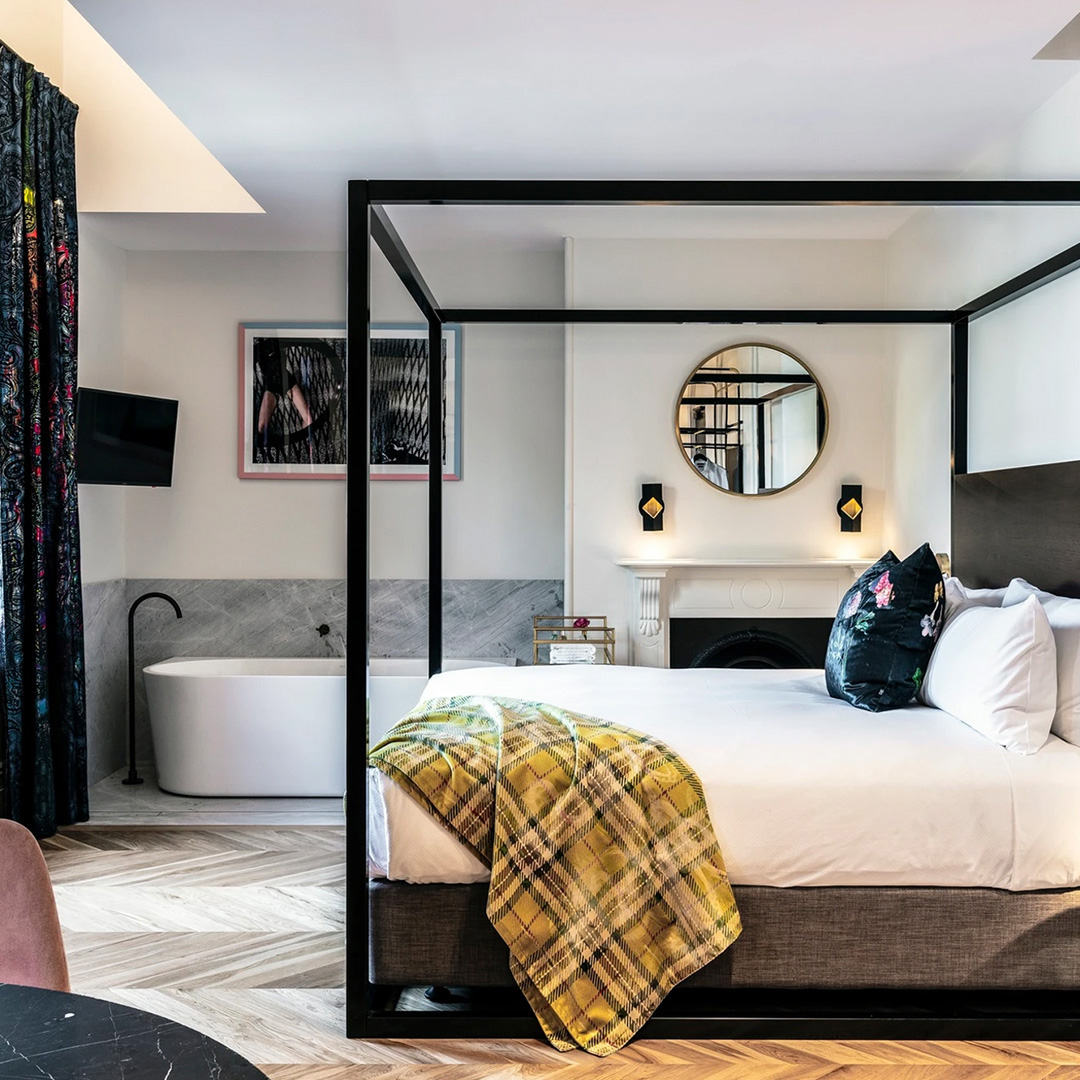 A stylish hotel room featuring a four-poster bed, freestanding bath, and herringbone flooring.