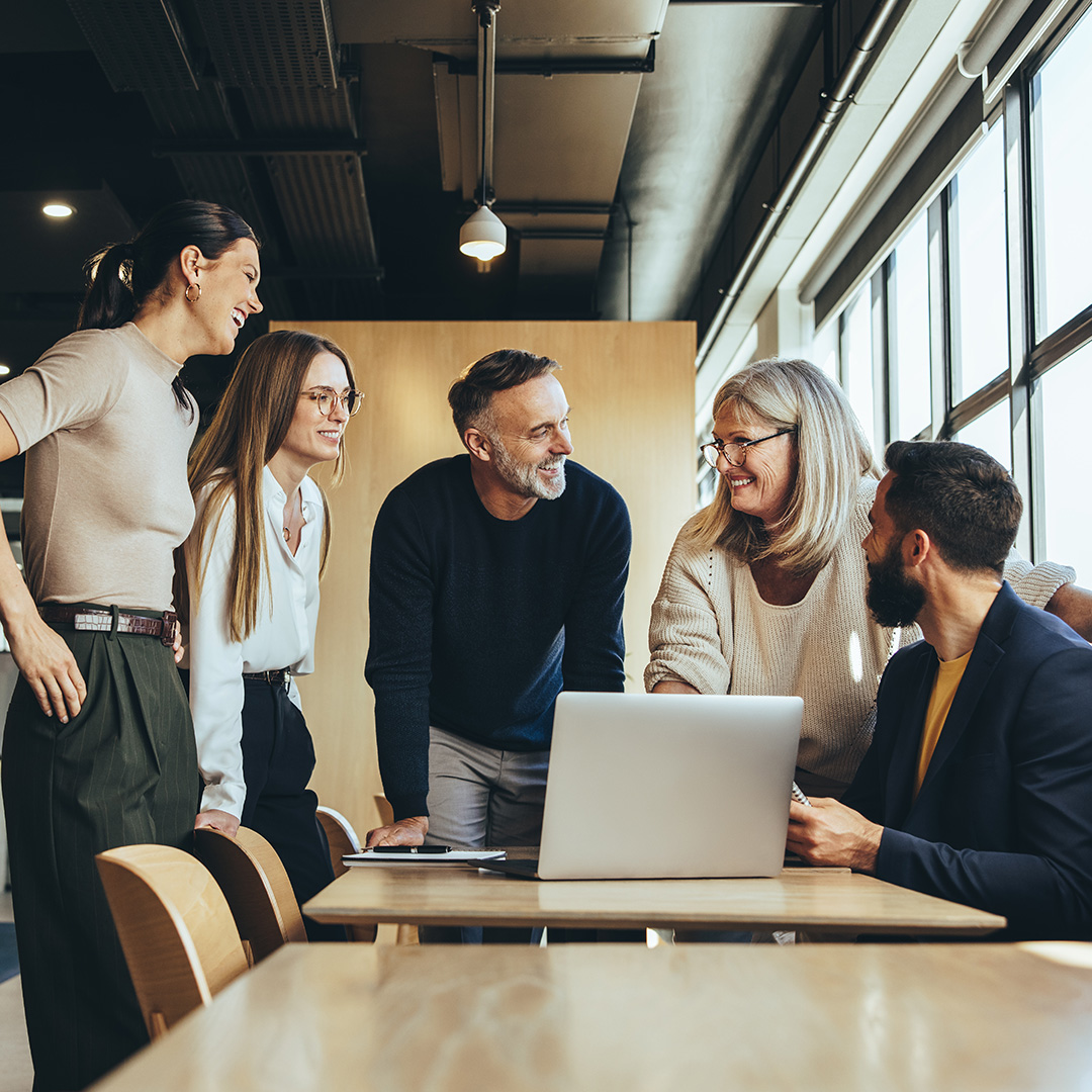 The image shows colleagues gathered around a table discussing a project in an open plan workspace.