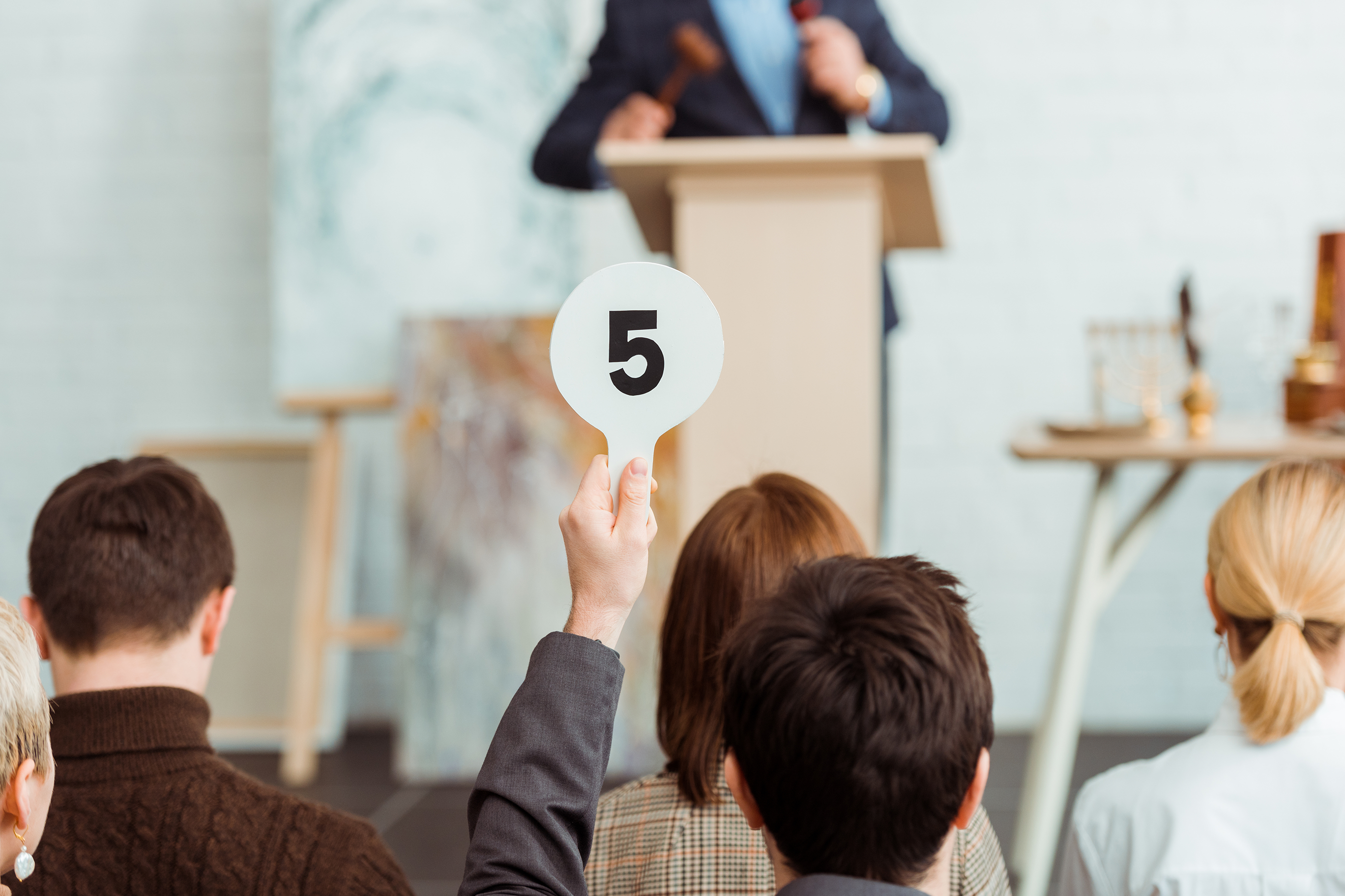 A person at an auction holds up a paddle with the number five on it while an auctioneer stands at a podium in the background.
