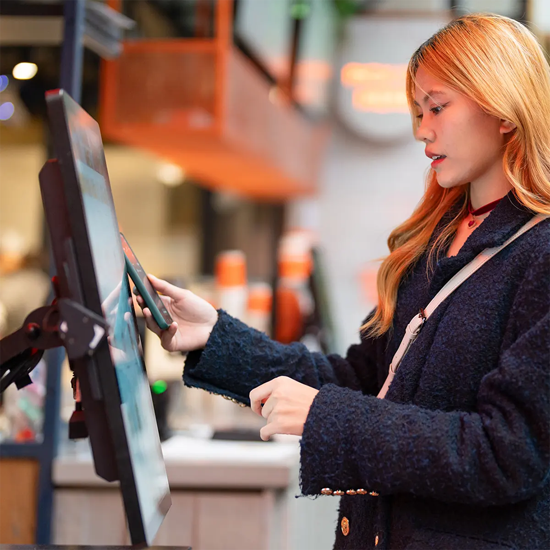 A young woman with blonde hair wearing a navy jacket uses her smartphone to make a contactless payment at a self-service kiosk.