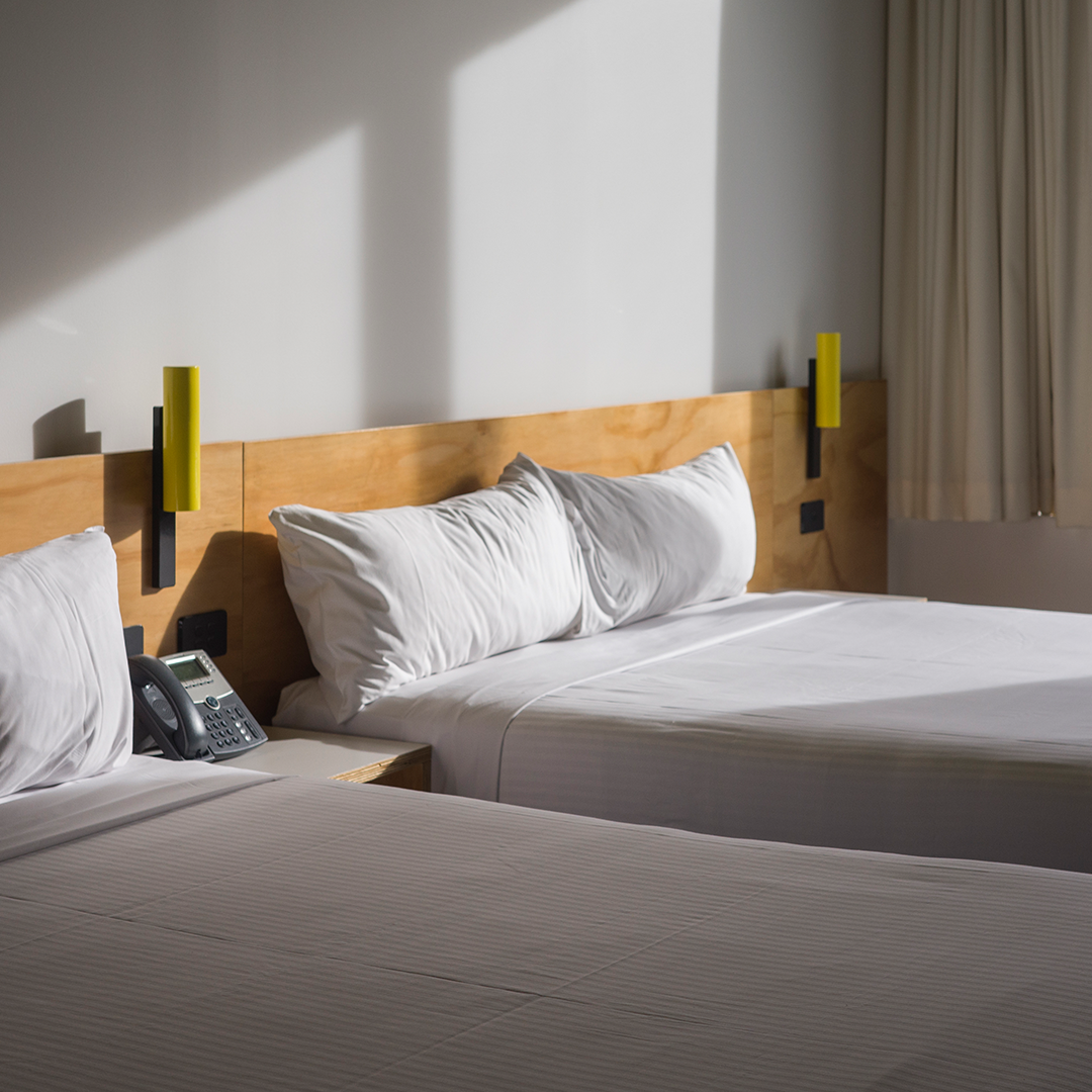 Two beds with white linen and pillows are shown in a modern hotel room with wooden headboards and yellow wall lights.