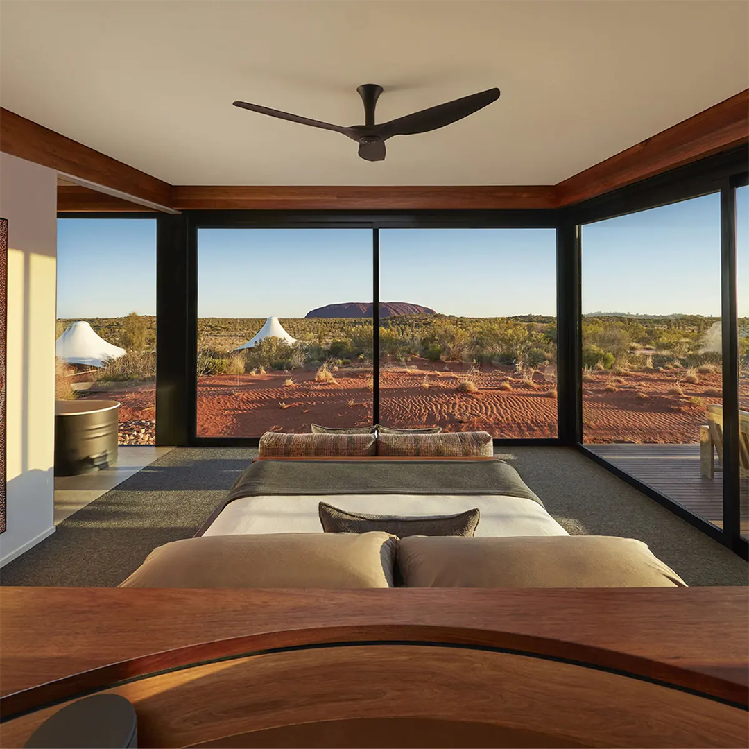 A luxurious bedroom with a king-sized bed offers a breathtaking panoramic view of the Australian Outback, featuring Uluru in the distance, through expansive floor-to-ceiling windows.