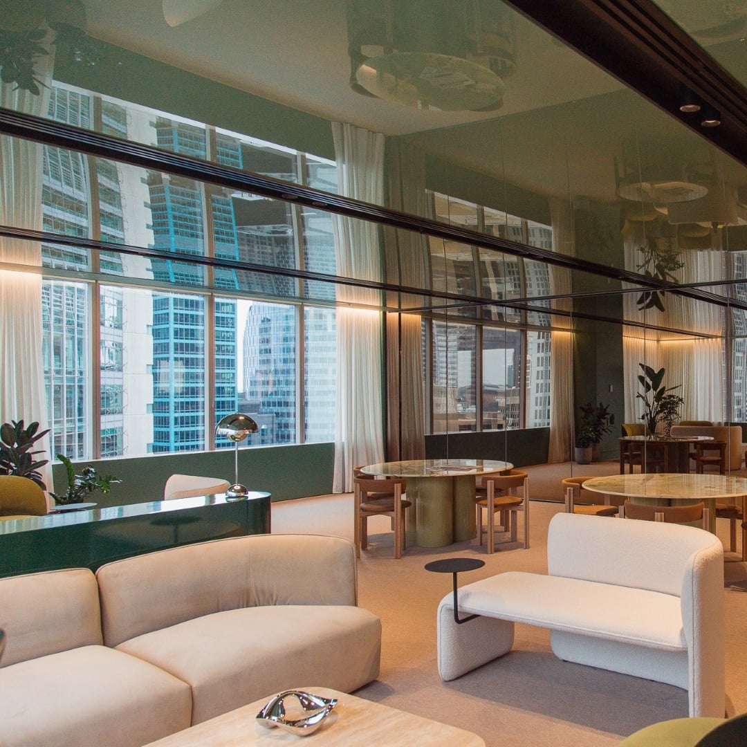 A sophisticated and modern lounge area at CBRE Sydney's office features plush beige and white seating, round dining tables, and expansive windows offering city views, all reflected in the mirrored ceiling.