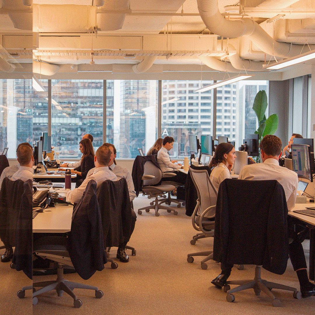 A bustling open-plan office features numerous employees working at desks with computers, set against a backdrop of a city skyline visible through large windows.