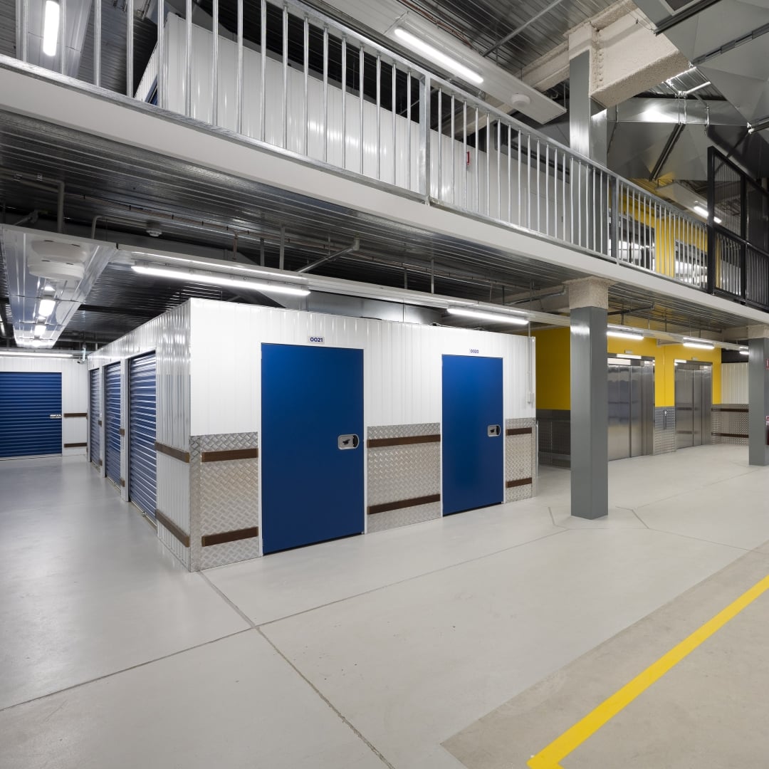 A brightly lit indoor self-storage facility features rows of blue-doored units, some with roller shutters, and a second-level walkway with white railings overlooking the ground floor.