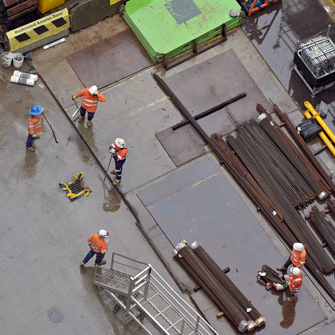 An aerial view shows several construction workers on a wet, grey concrete surface surrounded by stacks of metal rods, with some wearing high-visibility orange and blue gear.