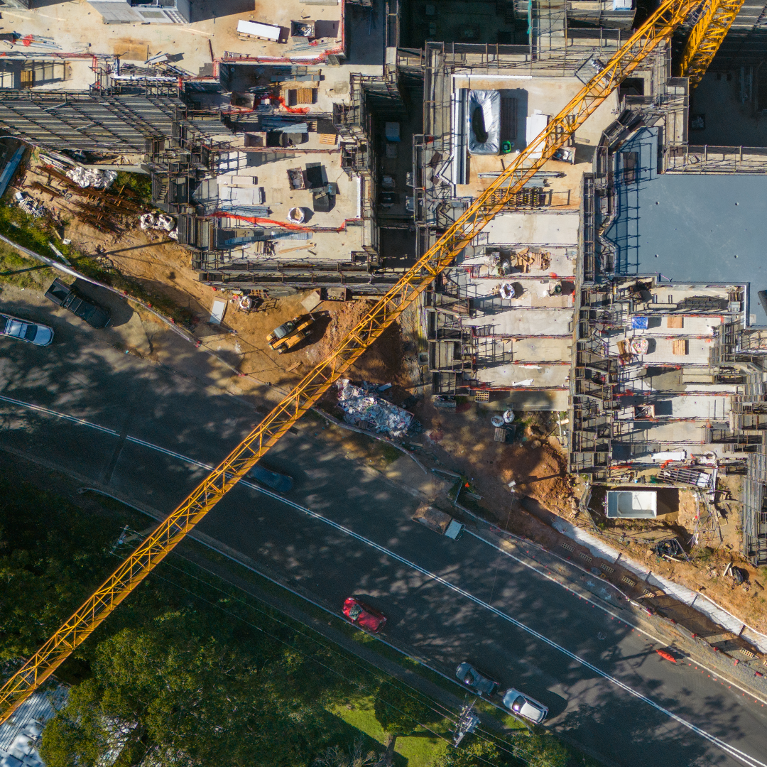 An aerial shot captures a large construction site with multiple unfinished buildings, a long yellow crane extending across a road with cars, and trees in the foreground.