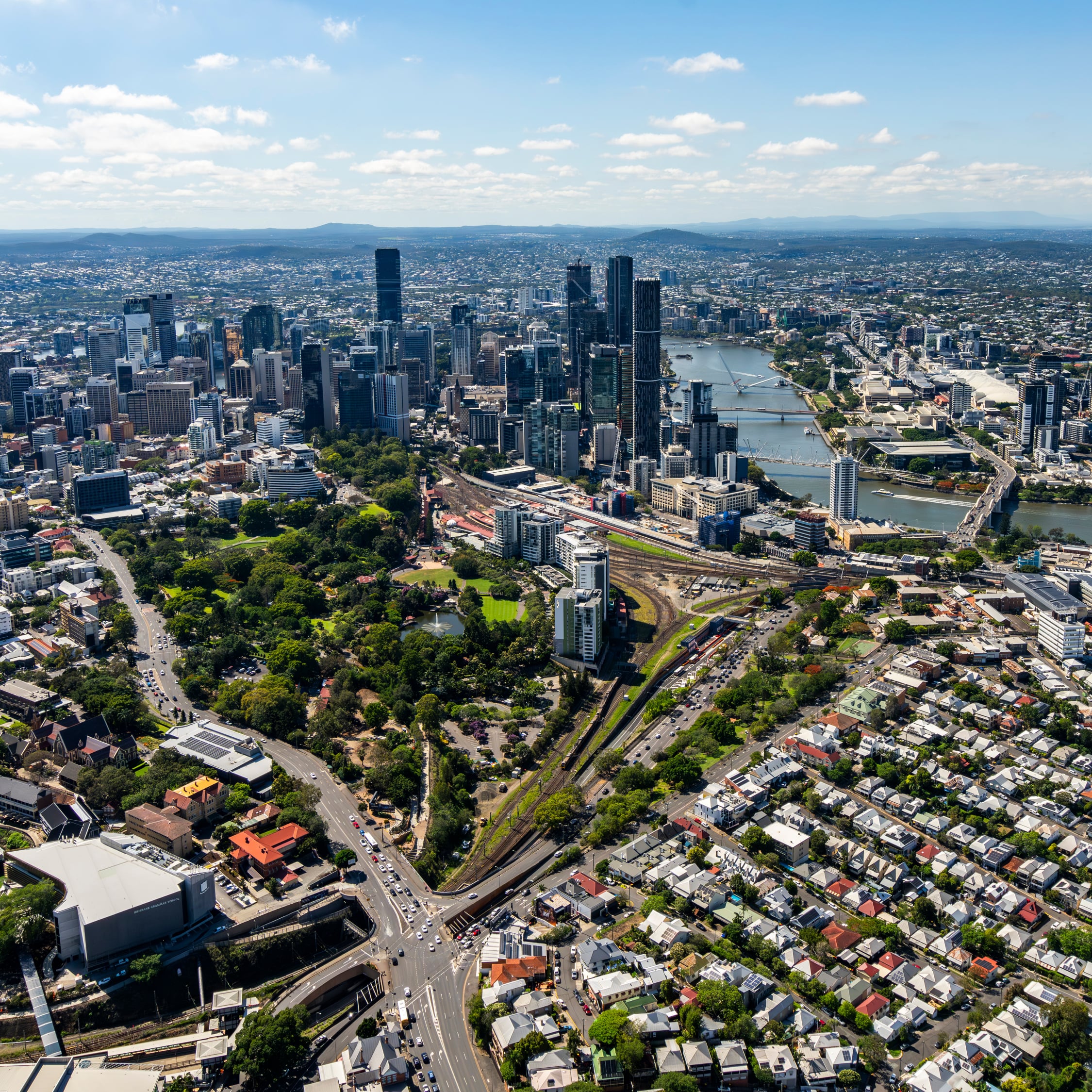 An aerial view of the Brisbane CBD featuring the winding Brisbane River, the lush greenery of Roma Street Parkland, and surrounding suburban residential areas. 