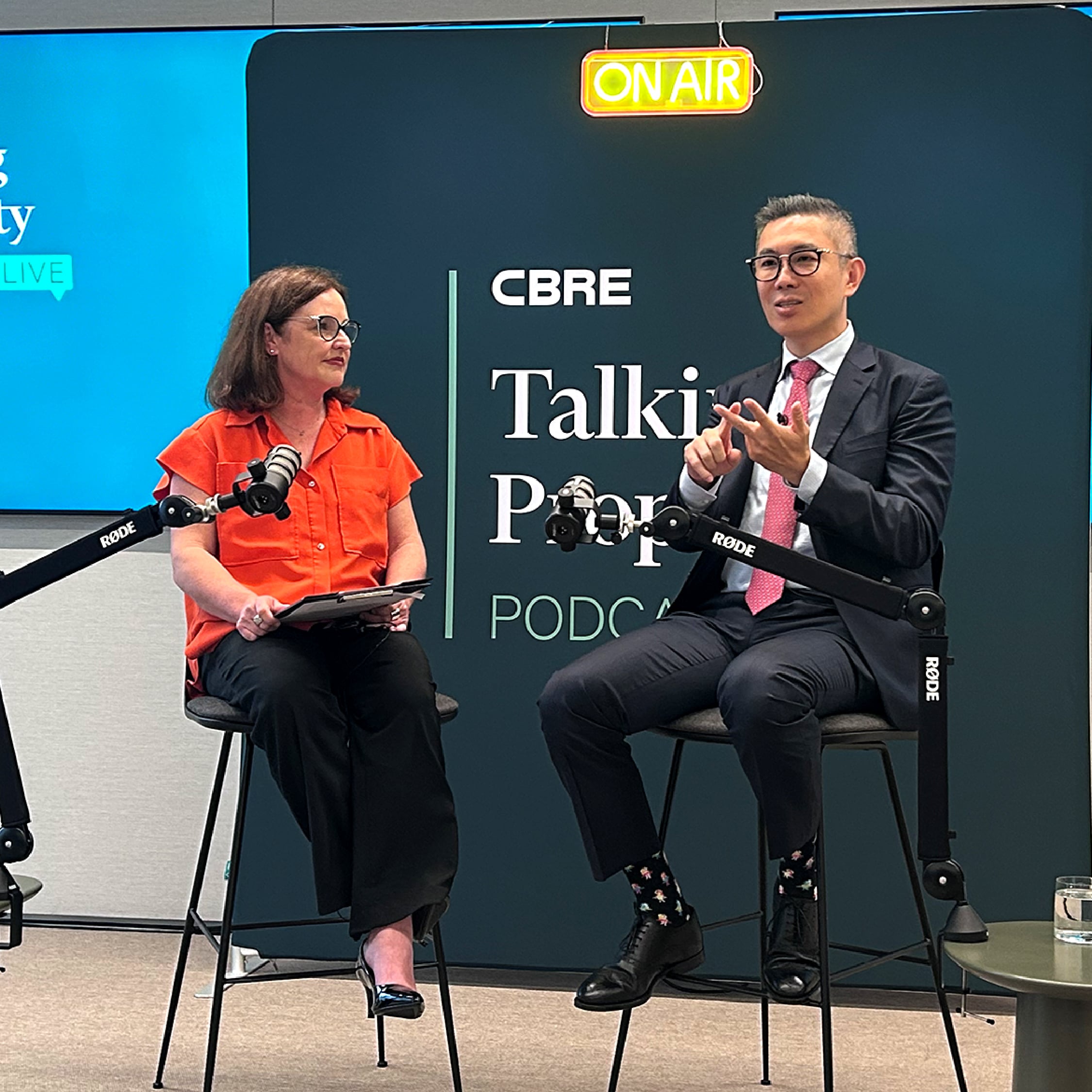Two presenters, Kathryn House and Henry Chin sit on high stools with microphones, recording a podcast in front of a dark green banner that reads "CBRE Talking Property Podcast" with an "ON AIR" sign above them.