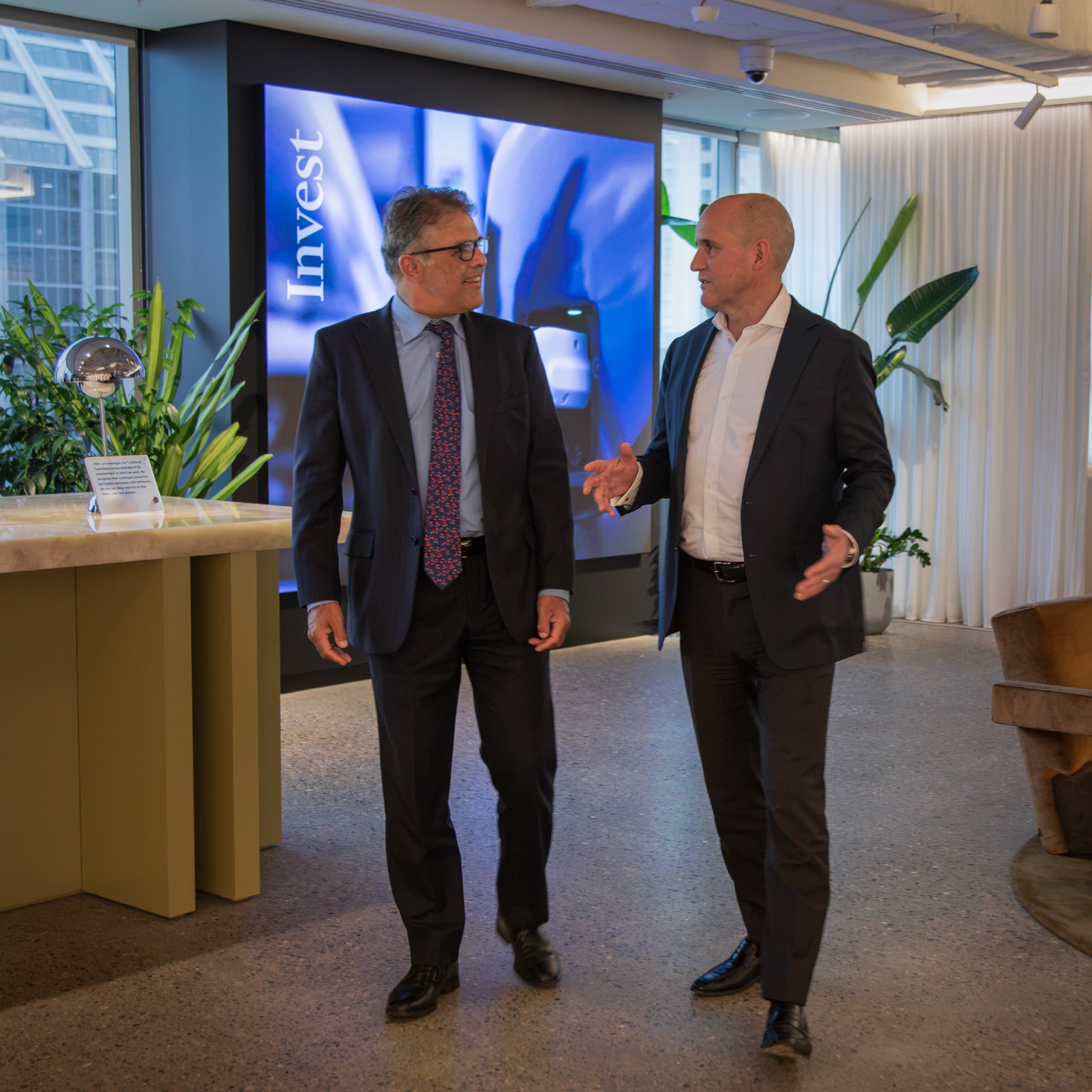 Sameer Chopra and Phil Rowland walk and talk together in a modern office lobby in front of a large digital screen.