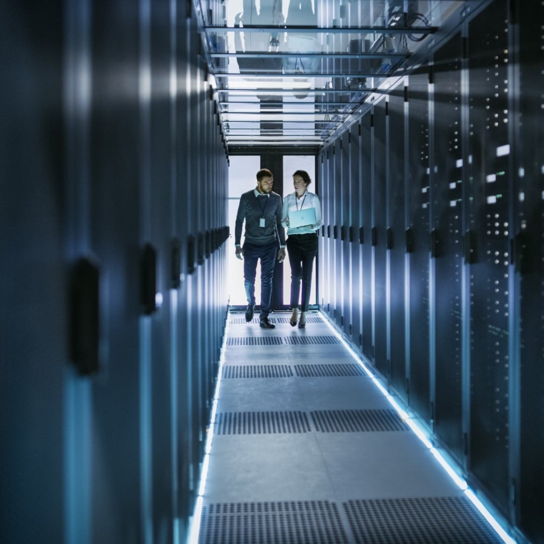Two professionals walk down a brightly lit aisle between rows of data servers in a modern data centre.
