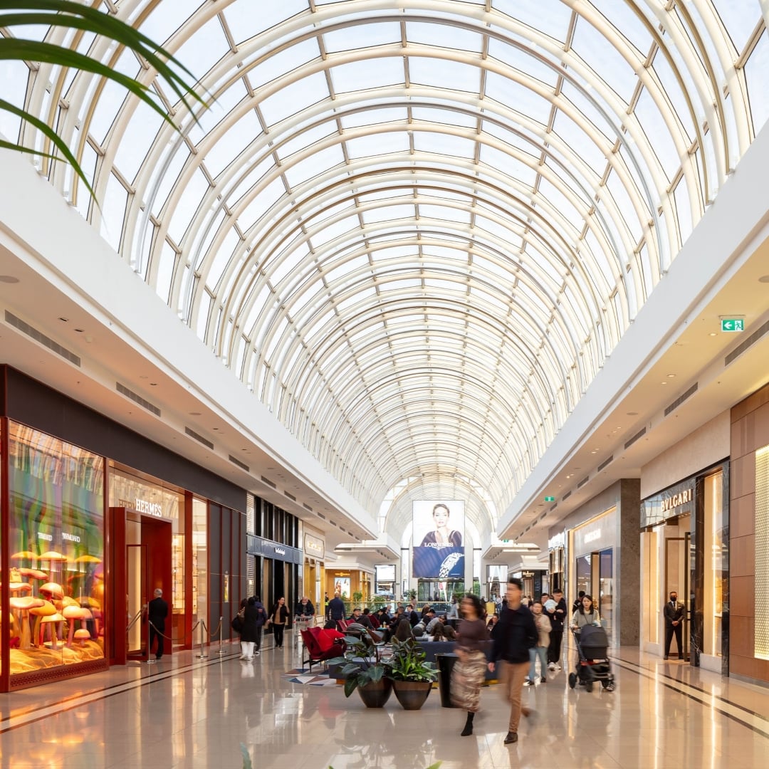 A brightly lit shopping centre features a vaulted glass ceiling, with luxury boutiques like Hermès and Bvlgari lining the polished floors.