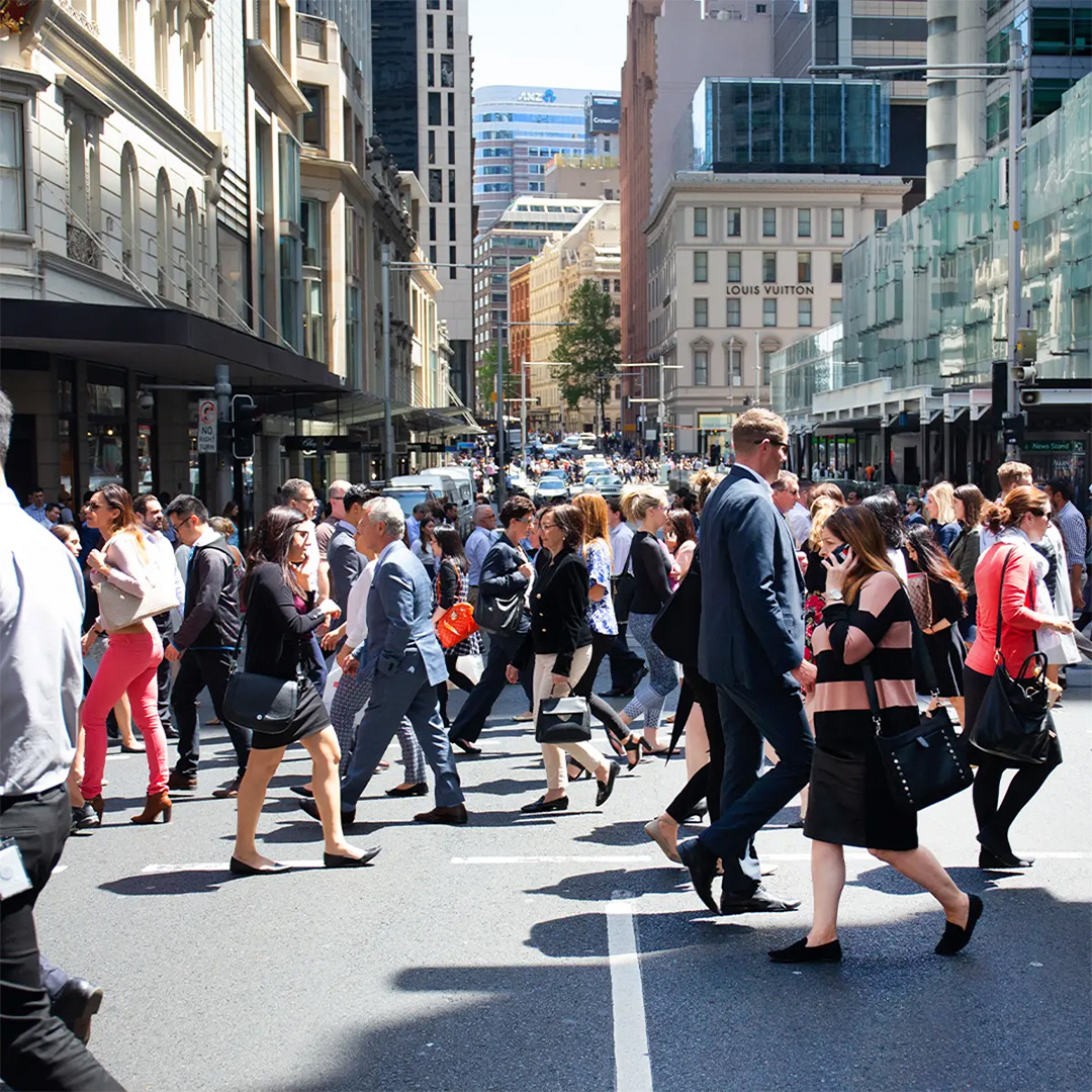 A bustling city street is filled with numerous pedestrians crossing a busy intersection, surrounded by tall buildings and a clear blue sky.