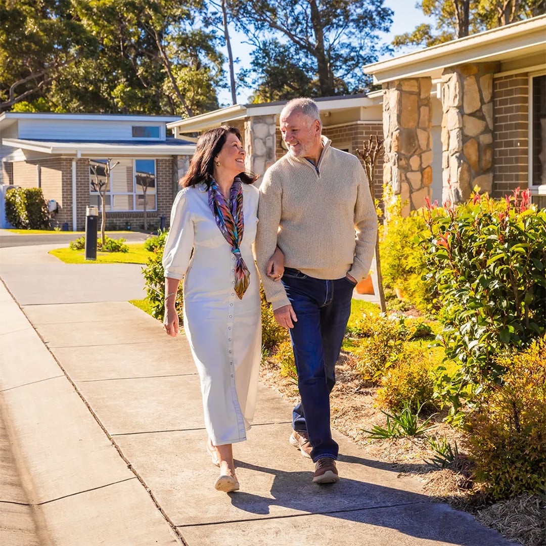 A happy older couple walks arm-in-arm along a sunny suburban footpath outside their home.