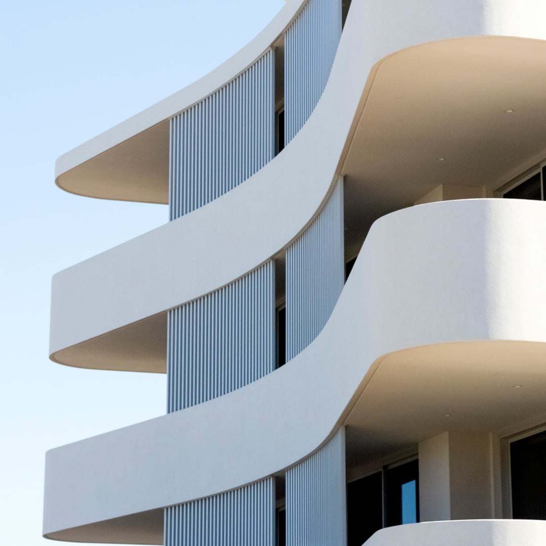 A modern apartment block featuring sleek, curved balconies and vertical metal screening against a clear blue sky.