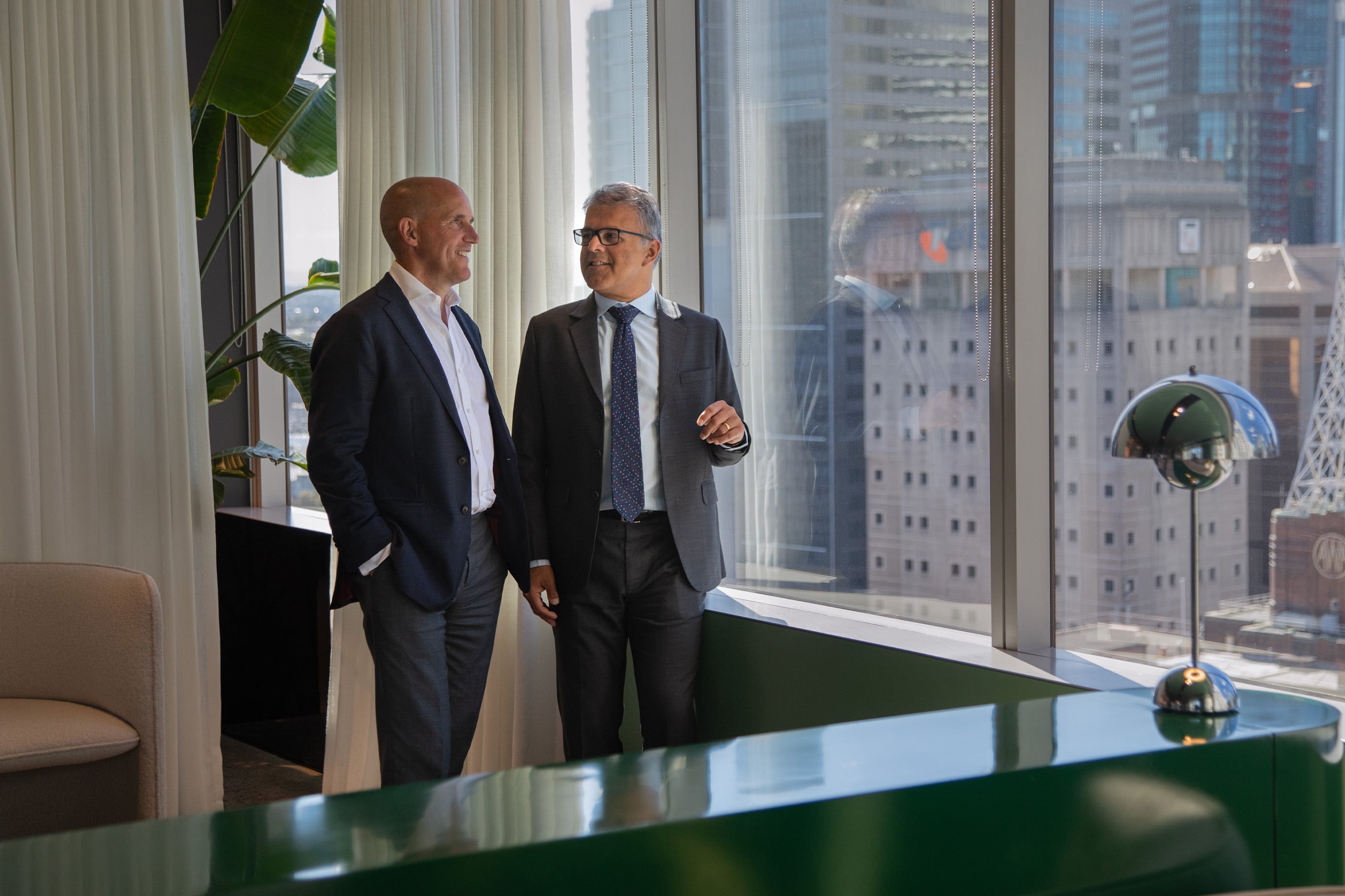 Phil Rowland and Sameer Chopra stand by a large office window, smiling and chatting while looking out over a city skyline.