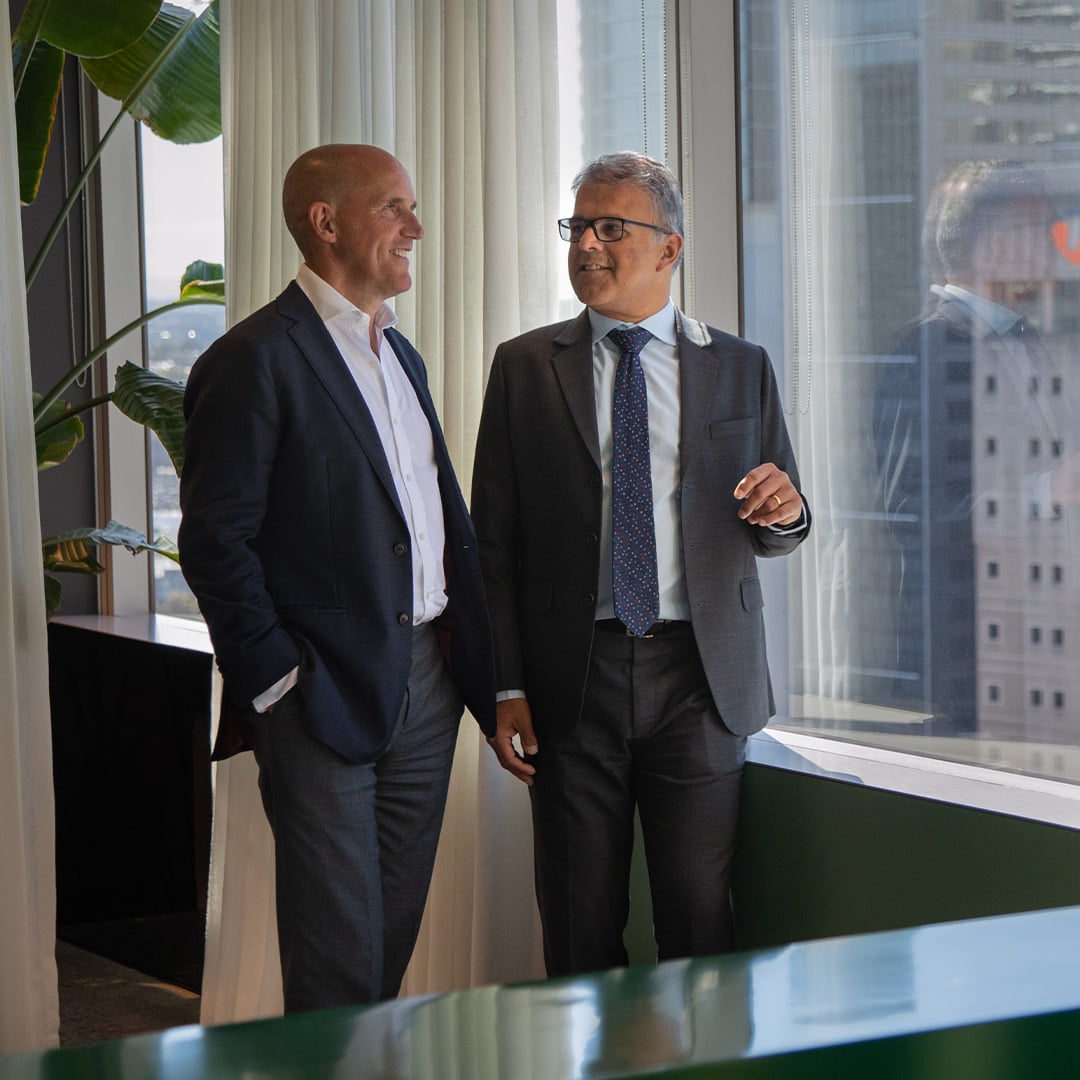 Phil Rowland and Sameer Chopra stand by a large office window, smiling and chatting while looking out over a city skyline.