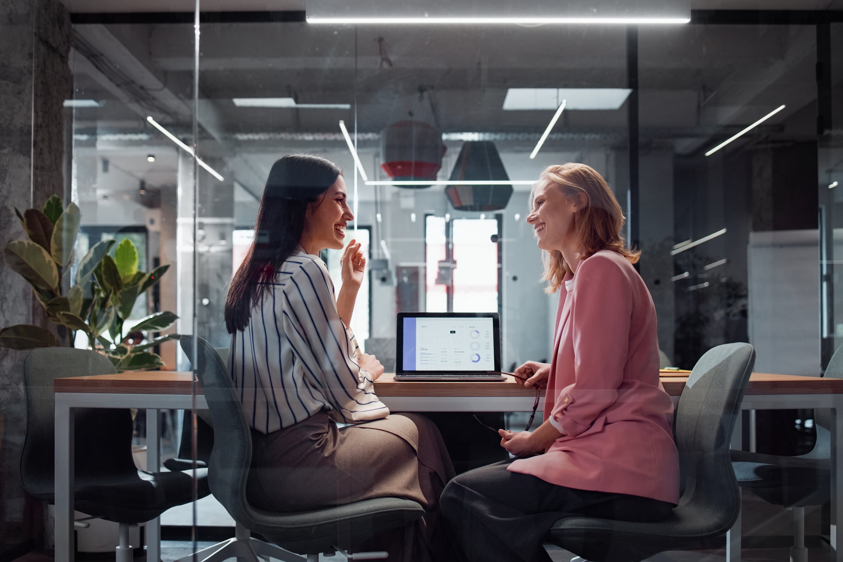 Two professional women sit opposite each other at a boardroom table, smiling and chatting while reviewing data on a laptop.