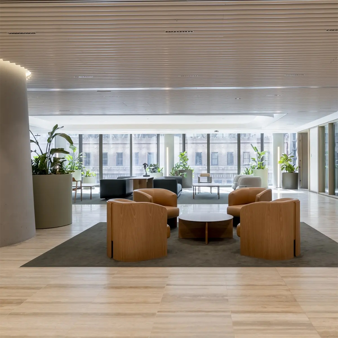 A modern office lounge featuring light timber armchairs arranged around coffee tables, set against a backdrop of floor-to-ceiling windows and potted plants.