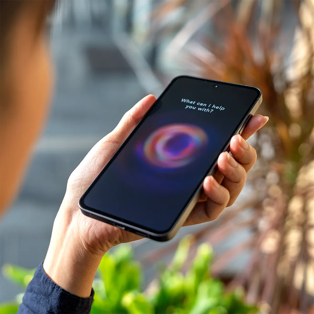 A person holds a smartphone displaying a colourful, glowing orb and the prompt "What can i help you with?" on its screen.