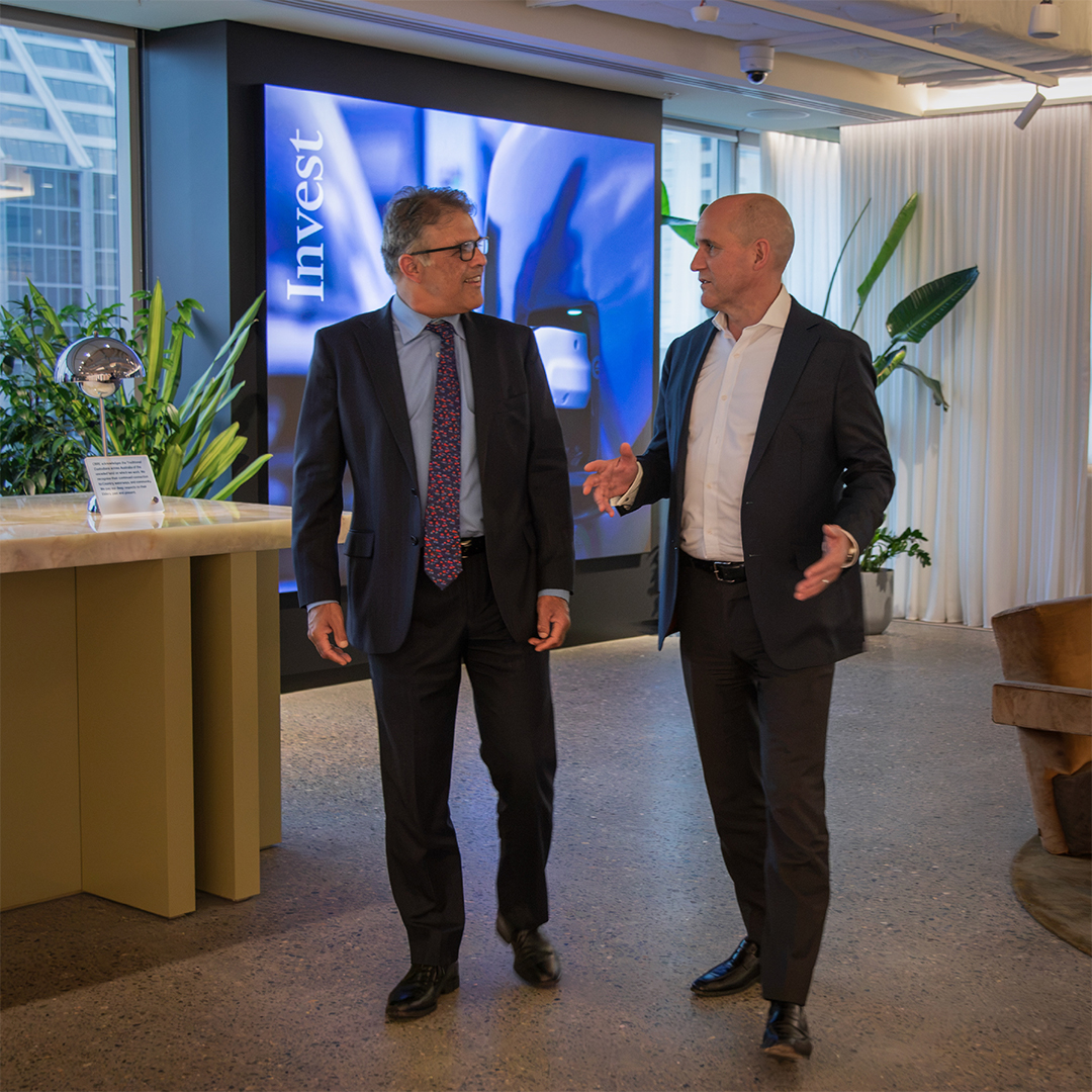 Sameer Chopra and Phil Rowland walk and talk together in a modern office lobby in front of a large digital screen.