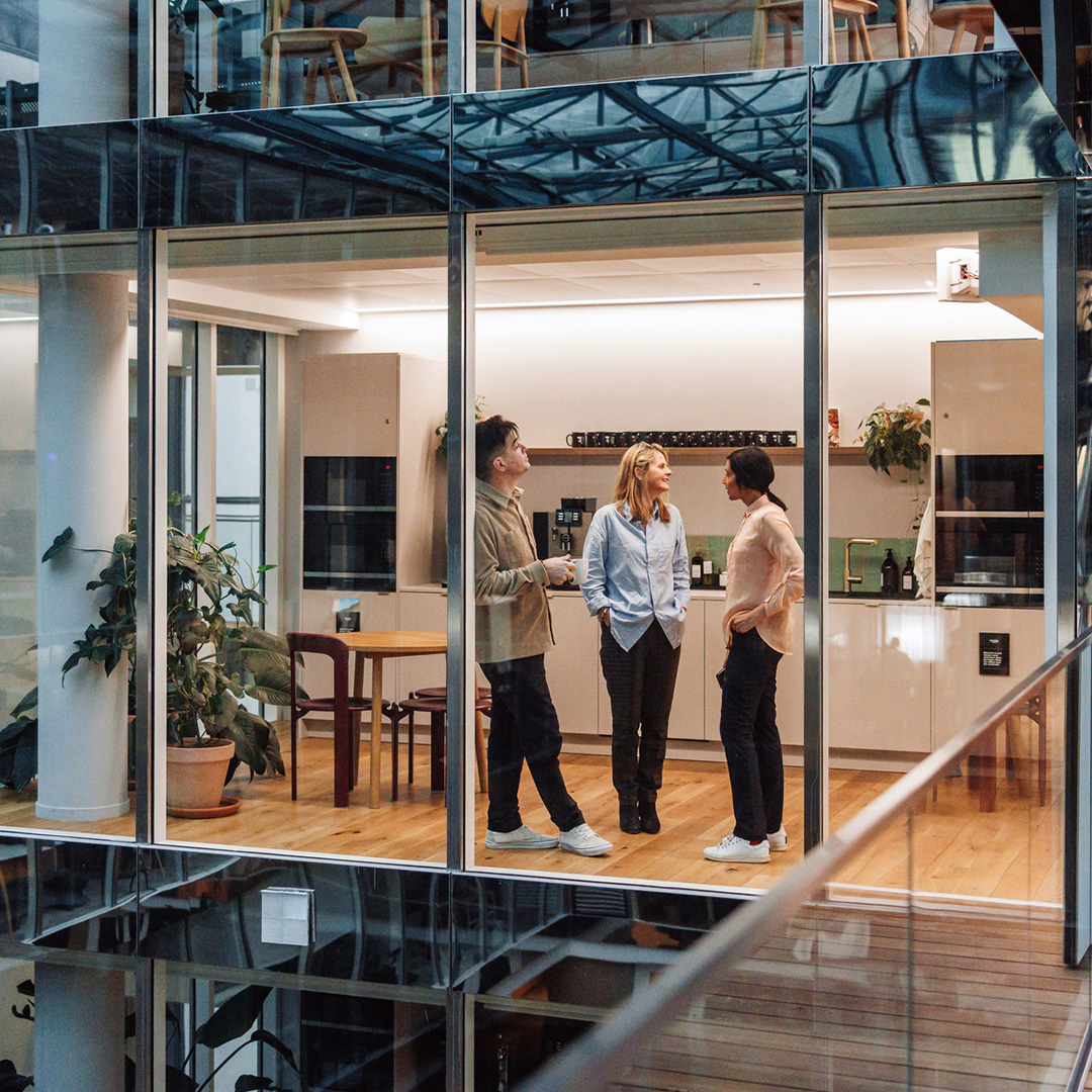 Three colleagues chat in a modern, glass-walled office kitchen featuring light wooden floors and minimalist cabinetry.