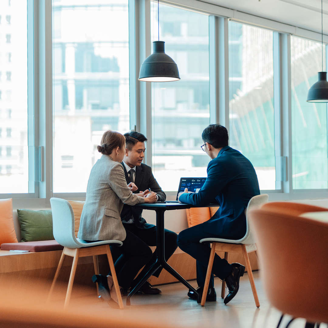 Three business professionals sit around a small table in a bright, modern office to discuss data on a laptop.