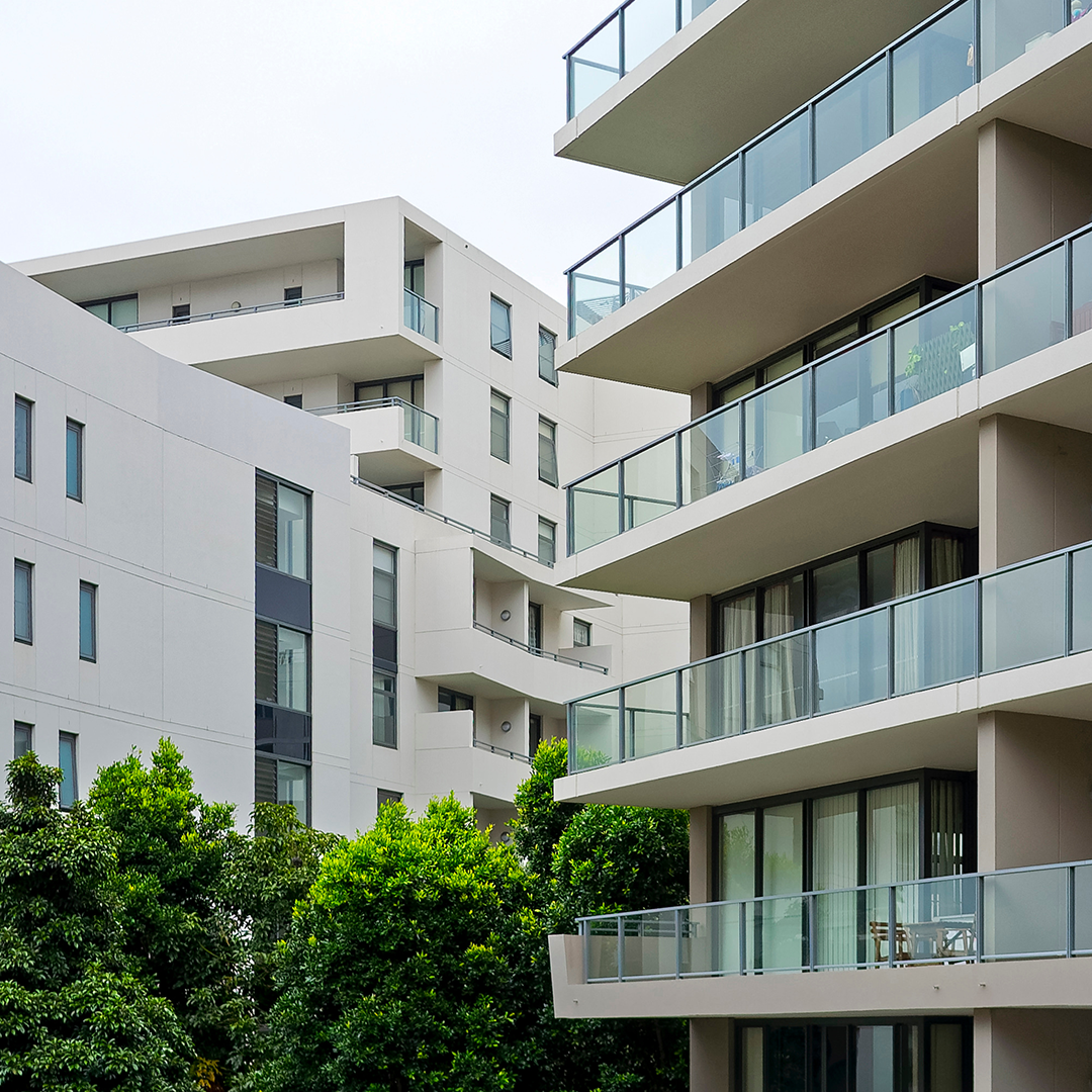A modern apartment building with multiple balconies featuring glass railings and lush green trees in the foreground.