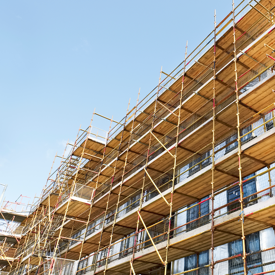 A building under construction is covered in extensive scaffolding, reaching upwards towards a clear blue sky.