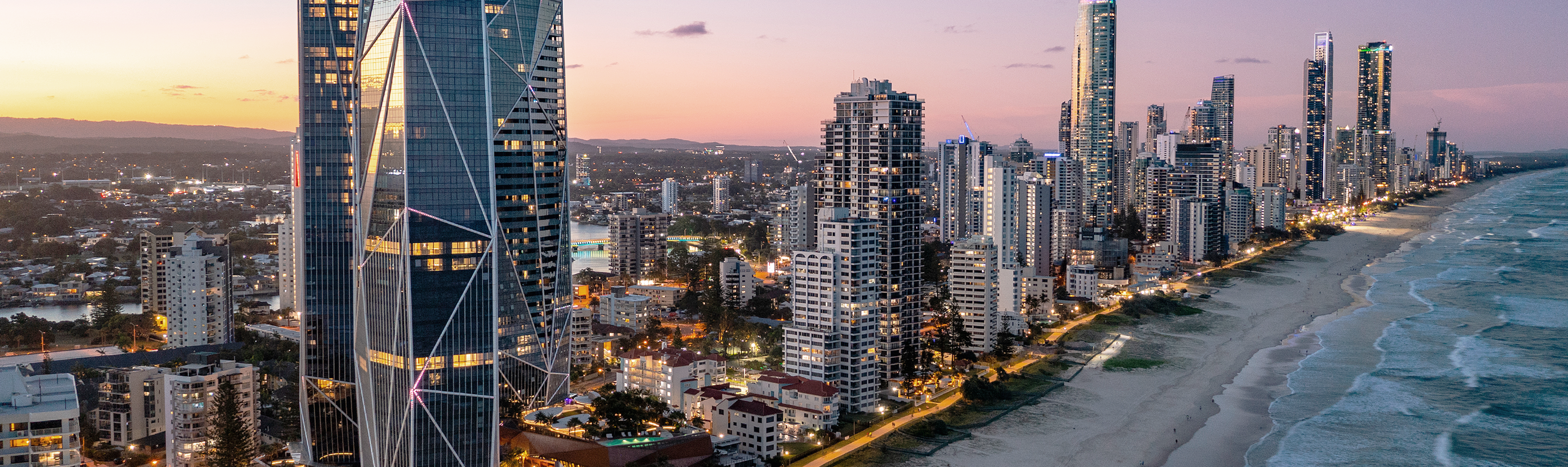 An aerial view of the Gold Coast skyline at dusk shows illuminated high-rise buildings lining a sandy beach with breaking waves, under a pink and orange sky.