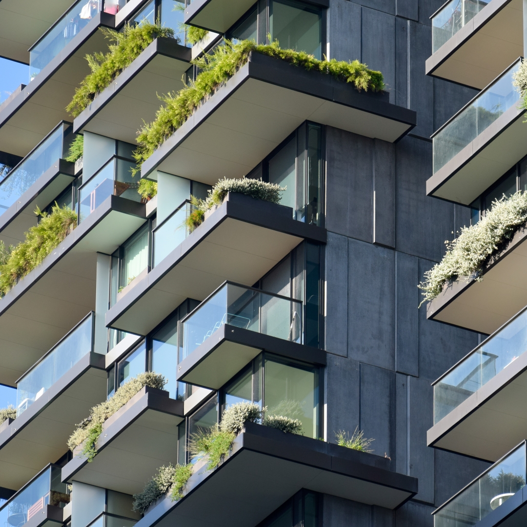 This close-up shot shows the modern architecture of a building with multiple balconies adorned with lush green and white plants, contrasting against the dark grey facade and clear glass railings.