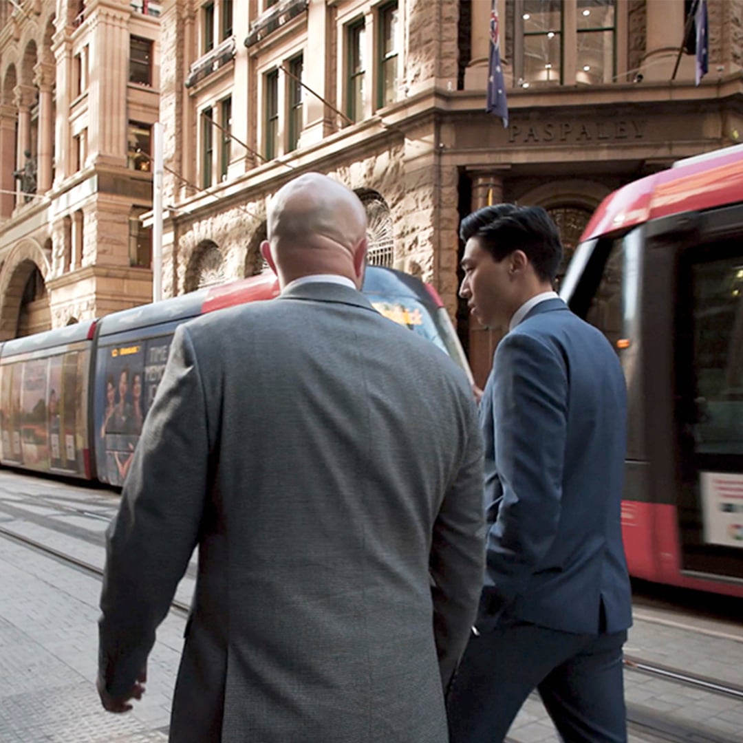 Two people in suits walk down George Street in Sydney, alongside a light rail tram.