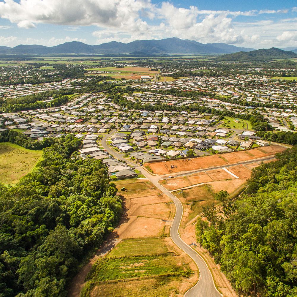 Aerial view of a large residential area with new housing developments and surrounding lush greenery, mountains in the distance.
