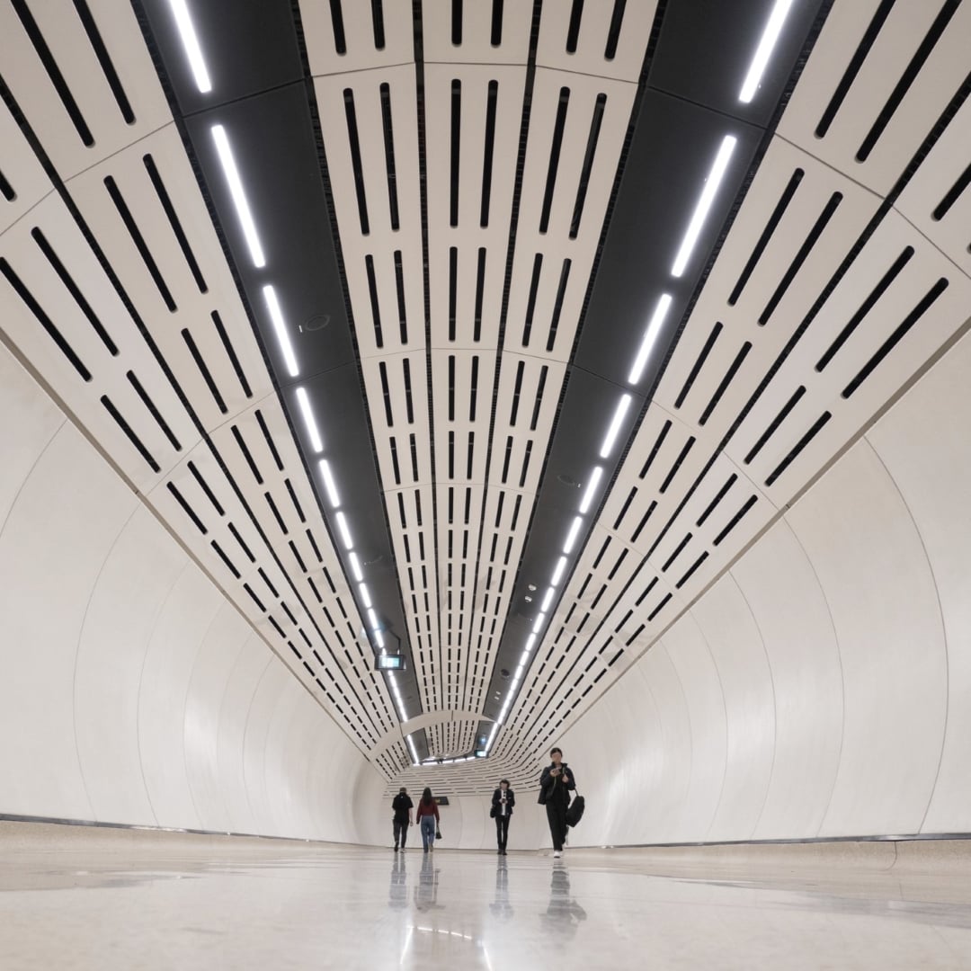A group of people walk through a modern, white-walled tunnel with a black and white patterned ceiling featuring strip lighting.