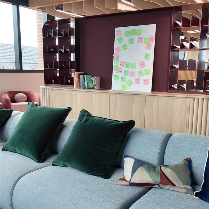 A light blue sofa with dark green throw pillows sits in front of a wall with a whiteboard covered in sticky notes, shelves, and a burgundy accent wall.