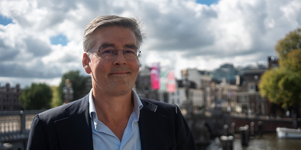 Man wearing glasses in front of the Stopera near Waterlooplein in Amsterdam. In the background, you can also see the Amsterdam canals and the Blauwbrug bridge.