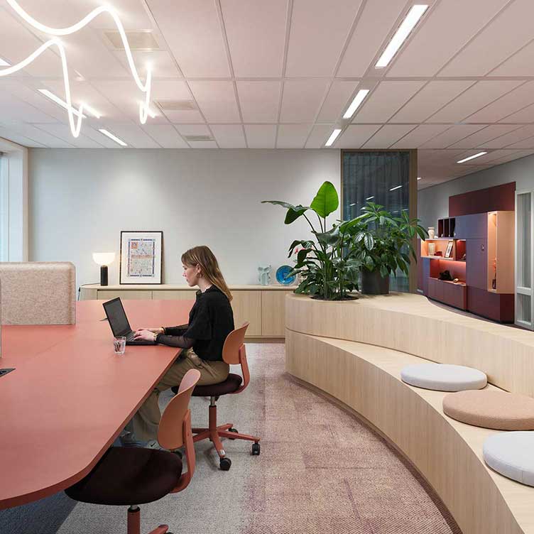 Moderne kantoorruimte met grote werktafel, ergonomische stoelen, planten en een gebogen zitbank in een lichte, open werkomgeving.