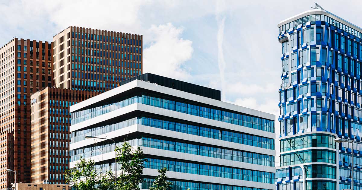 A cityscape with several modern buildings under a partly cloudy sky, showing contrasting architectural designs including brown lattice patterns, white stripes and blue glass.