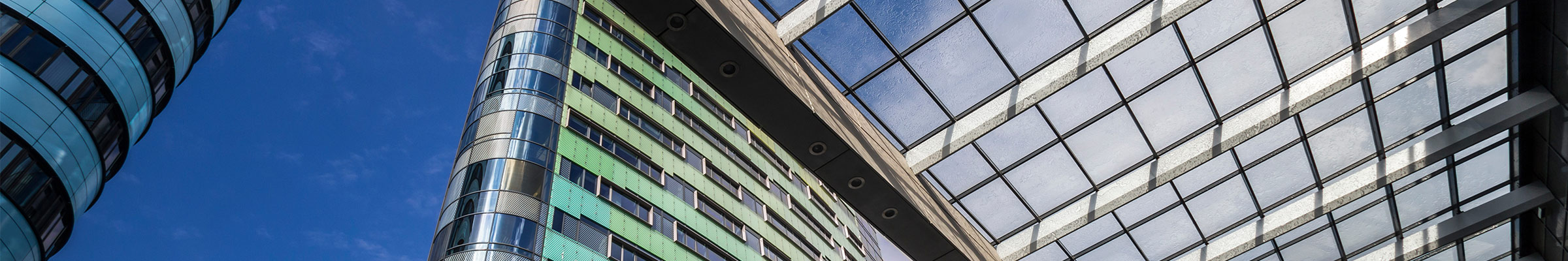 Een architecturale scène met de nadruk op wolkenkrabbers en glazen structuren. De lucht is helder met weinig wolken, en het perspectief kijkt omhoog naar de toppen van de gebouwen. De glazen structuur lijkt deel uit te maken van een plafond of overkapping, wat interessante geometrische patronen tegen de lucht creëert. Het gebouw aan de linkerkant heeft een cilindrische vorm met reflecterende ramen, terwijl het gebouw in het midden groenachtig glas heeft en meer rechthoekig van ontwerp is.