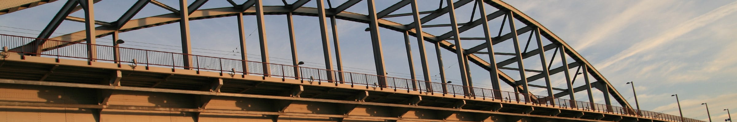 Een grote, gebogen en metalen brug die over een rivier loopt. De lucht is helder met een kleurverloop van blauw naar warme tinten, wat suggereert dat het ofwel zonsopgang of zonsondergang is. De lichtomstandigheden creëren een interessant contrast en benadrukken de structurele details van de brug.