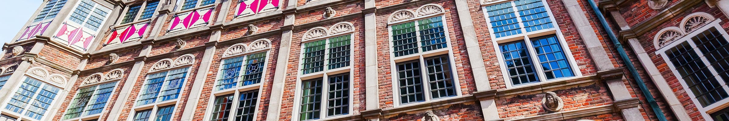 De gevel van een historisch gebouw met sierlijke architectuur. Het gebouw heeft meerdere rijen hoge, rechthoekige ramen met witte kozijnen, geplaatst in een rode bakstenen muur. Boven de ramen hangen decoratieve witte en roze banners tussen stenen beeldhouwwerken en sculpturen. De daklijn is versierd met een reeks kleine ramen of dakkapellen, ook omlijst in wit. De lucht is helder en blauw, wat wijst op goed weer en de details en kleuren van het gebouw benadrukt.