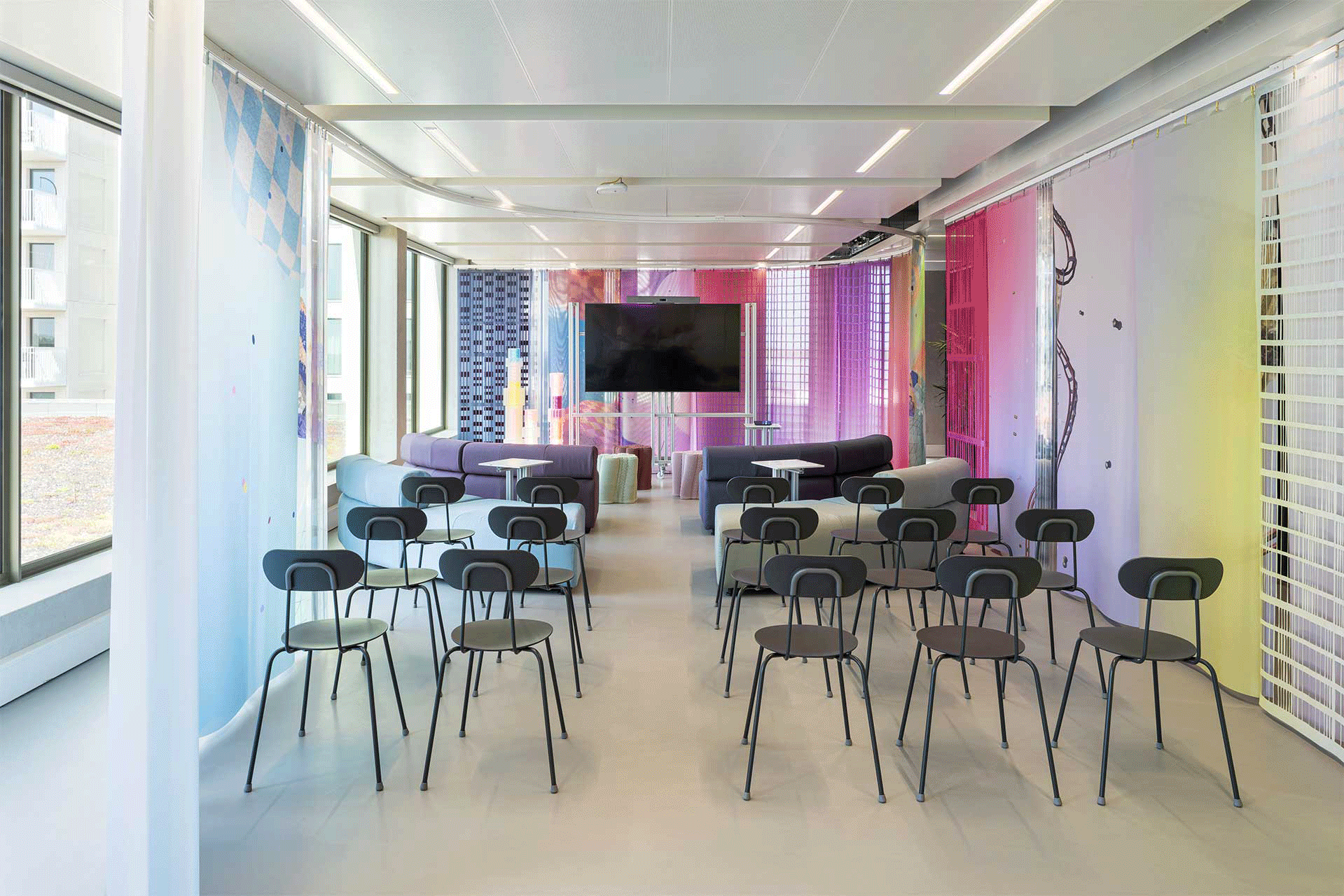 A meeting room with rows of chairs and benches, a large presentation screen, and colorful wall panels, suitable for presentations, workshops, and meetings in a bright office setting.