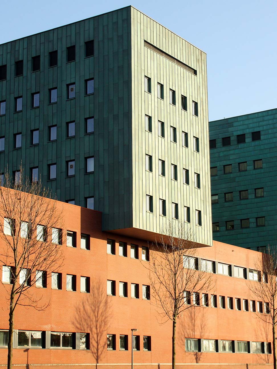 Groot kantoorgebouw of zorgcomplex met rode bakstenen gevel en groene torens, omringd door bomen onder een heldere blauwe lucht in stedelijke omgeving.