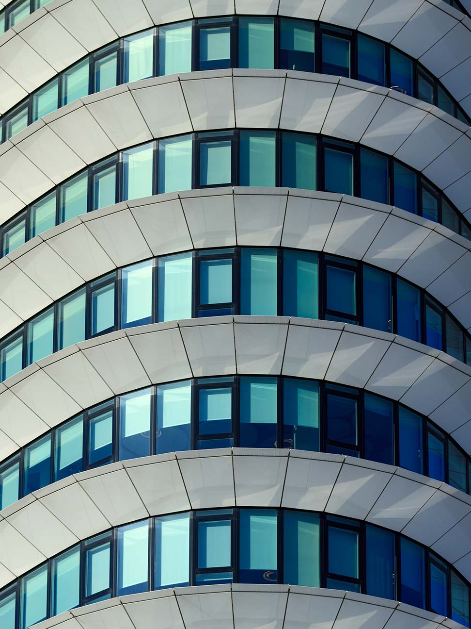 Afgeronde gevel van hoog kantoorgebouw met blauwe glaspartijen en witte gevelpanelen in gelaagd patroon, geschikt voor commercieel vastgoed en architectuur.